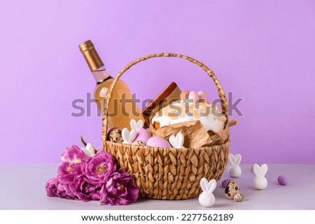 Basket with Easter eggs, cake, bottle of wine and tulip flowers on lilac background