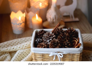 Basket with pine cones and seasonal spices on the table. Lit candles in the background. Hygge at home. Selective focus. - Powered by Shutterstock