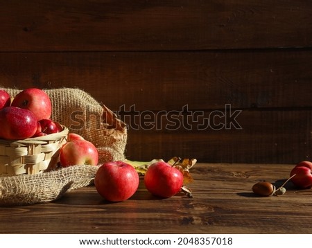 Basket with red apples on sackcloth close up. Dry oak leaves, acorns, ripe fruits on wooden table at brown wood barn wall background. Autumn food, fall menu, thanksgiving, harvest season concept