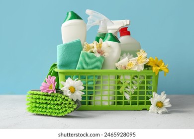 Basket with set of cleaning supplies and spring flowers on white table against blue background - Powered by Shutterstock