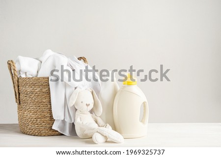 A basket of white laundry, a teddy bunny toy, a bottle of liquid detergent, washing gel or fabric softener. Mockup for washing baby clothes with copy space.