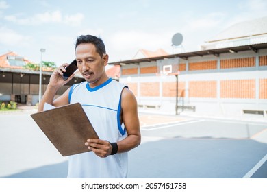 A Basketball Coach Calls Using A Mobile Phone While Holding A Clipboard