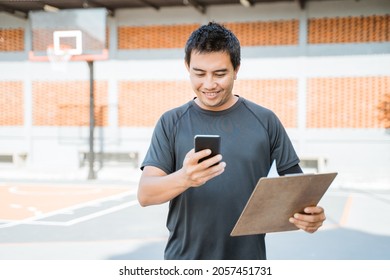 Basketball Coach Looking At A Mobile Phone Screen While Holding A Clipboard