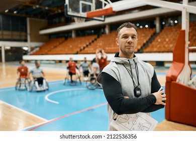 Basketball coach standing with his arms crossed and looking at camera while his team in wheelchairs while practicing in the background. - Powered by Shutterstock