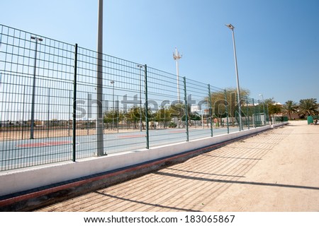 a basketball court fence against a blue sky with a sand path in the foreground