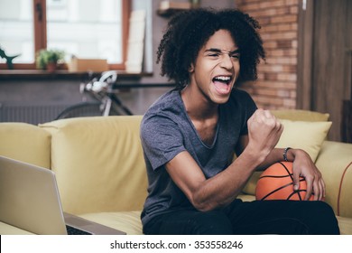 Basketball Fan. Cheerful Young African Man Watching TV And Holding Basketball Ball While Gesturing On The Couch At Home