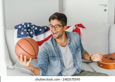 Basketball Fan. Cheerful Young Man Watching TV And Holding Basketball Ball While Gesturing On The Couch At Home. Man Watching Basketball Game On Tv And Celebrating Victory, American Flag Behind Him.