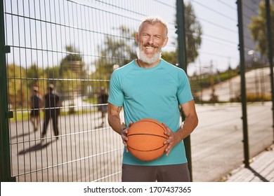 Basketball player. Happy mature man in sportswear holding basketball ball and smiling at camera while standing at outdoor basketball court - Powered by Shutterstock