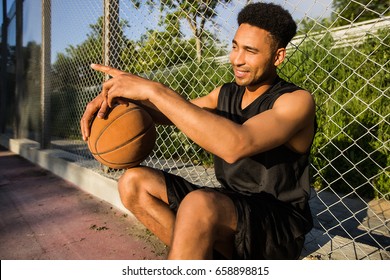 Basketball player is sitting on a basketball court,man with a ball.sport outfit,sport competitions,sportsman,black shorts,game,black t-shirt,bracelets,happy,attractive,laugh,sneakers,athlete,active