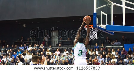 A basketball player throws a ball into the hoop. In the background, the fans are sitting in the stands
