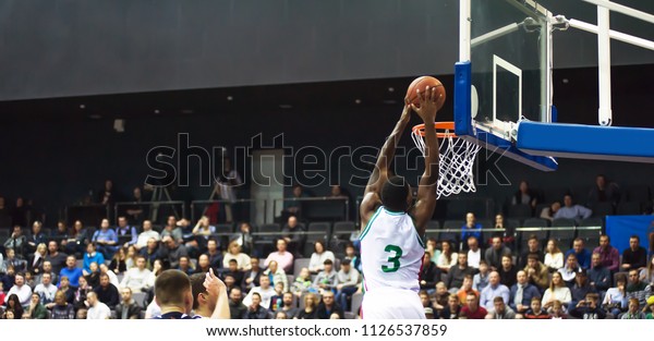 A basketball player\
throws a ball into the hoop. In the background, the fans are\
sitting in the stands