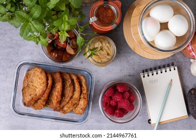 Batch Cooking Containers Full Of Homemade Meal, Eggs, Meat, Raspberries, Onion, Around A Spiral Notebook With The Word MENU Written Over A Cement-like Table.