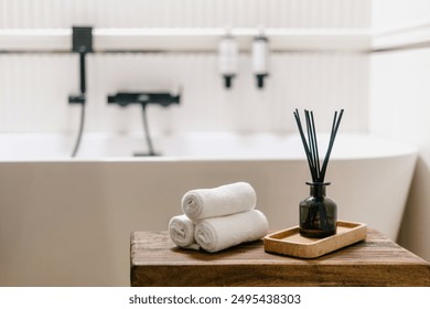 Bath accessories. Selective focus on wooden shelf with aromatic oil fragrance in glass bottle on tray close to rolled face towels against blurred bathroom interior. Concept of home spa and treatment