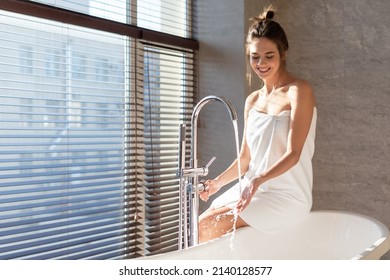 Bath Time. Attractive Millennial Woman Wrapped In Towel Sitting On Bathtub And Playing With Water Flowing From Tap, Young Beautiful Lady Spending Time In Modern Bathroom At Home, Copy Space