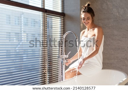 Bath Time. Attractive Millennial Woman Wrapped In Towel Sitting On Bathtub And Playing With Water Flowing From Tap, Young Beautiful Lady Spending Time In Modern Bathroom At Home, Copy Space
