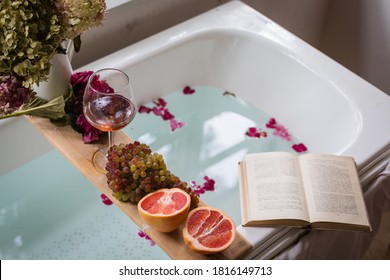 Bath tub with flower petals, grapefruit slices, bunch of grapes, a glass of wine, opened book and hydrangea bouquet. Organic spa relaxation preparation - Shutterstock ID 1816149713