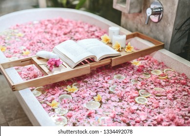 Bath Tub With Flower Petals And Lemon Slices. Book And Candles On A Tray. Organic Spa Relaxation In Luxury Bali Outdoor Bathroom.