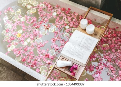 Bath Tub With Flower Petals And Lemon Slices. Book, Candles And Beauty Product On A Tray. Organic Spa Relaxation In Luxury Bali Outdoor Bathroom.