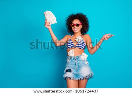 I bathe in money! Funny beautiful wealthy attractive african woman wearing pink glasses striped bra jeans skirt is playing with cash, isolated on blue background