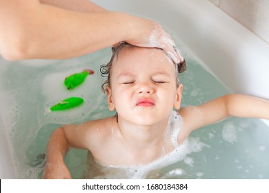 Bathing Without Tears. Patient Baby With Narrowed Eyes Takes A Bath And Washes His Head. Mom Washes The Babys Head With Shampoo.