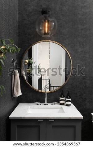 A bathroom with black snake skin wallpaper, circular gold mirror, marble countertop on a grey cabinet, and modern black light fixture.