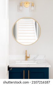 A Bathroom With A Blue Vanity Cabinet, Marble Countertop, And A Gold Faucet, Light Fixture, And Circular Mirror.