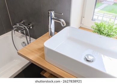Bathroom close-up: White rectangular sink with chrome faucet on a wooden countertop, next to a bathtub and window with greenery