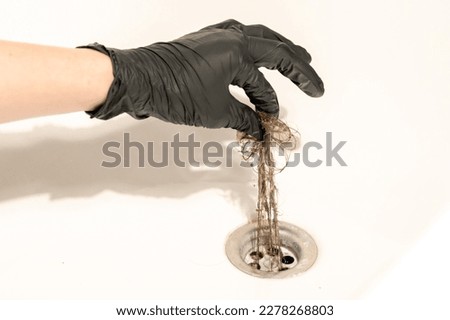 A bathroom drain clogged with dirty hair and slime. A female hand in a black latex glove pulls a long messy tuft of hair out of the drain