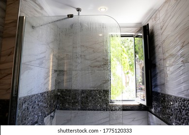 Bathroom Filled With Natural Light Through An Open Window. Shower Head Mounted To The Ceiling With Running Water. Modern Framless Fixed Glass Panel, Marble Textured Tile. Copy Space, Background.