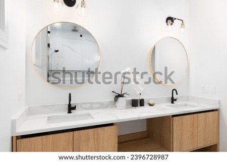A bathroom with a floating white oak cabinet, white marble countertop, and black and gold lights over gold circular mirrors.