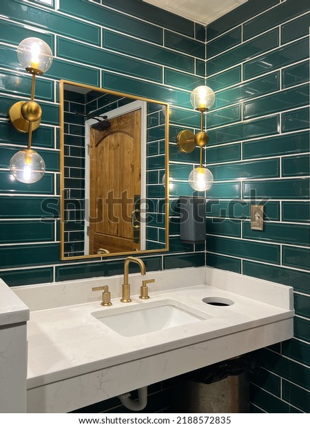 A bathroom with green subway tile walls,\
gold lights, marble sink and a gold\
faucet.