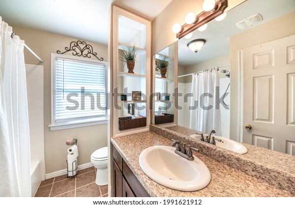 Bathroom interior with window, vanity sink and wood\
shelves divider with decorative display. There is a sink with\
granite top beside the toilet with toilet paper stand against the\
window near the tub.