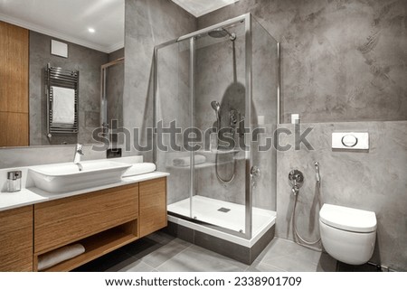 bathroom with luxury interior, glass shower cabin, wc bowl with concealed cistern tank, ceramic washbasin with faucet and wooden under sink cupboard with clean towel