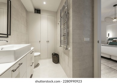 Bathroom with a mirror with a black metal frame, a white porcelain sink on a chest of drawers, a chrome radiator to dry towels and a gray built-in wardrobe - Powered by Shutterstock