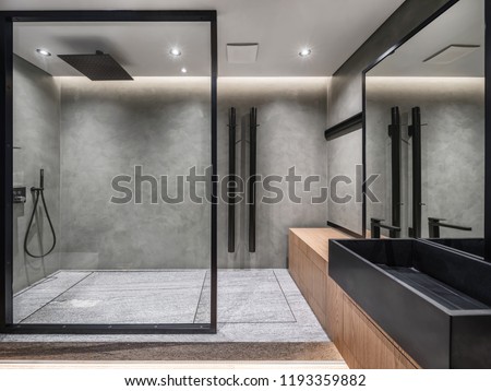 Bathroom in a modern style with gray tiled walls. There is a shower with a glass partition, wooden stand with a black sink and a faucet, large mirror, luminous lamps. Horizontal.