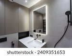 Bathroom in a modern style with gray and white tiles. There is a large mirror with luminous lamps, tabletop with wooden drawers and sink, bath with shower and glass partition, towel rack and a hanger.