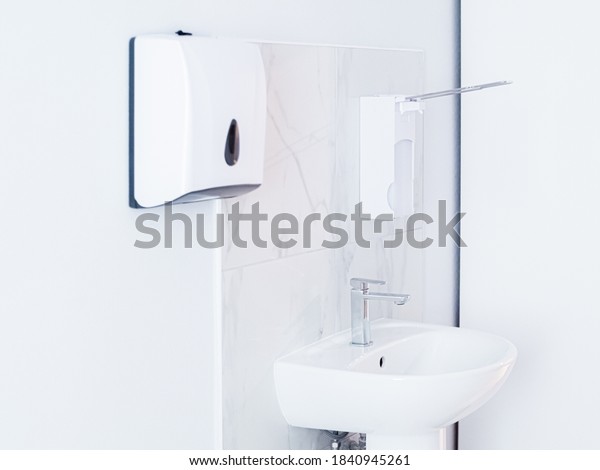 Bathroom with soap dispenser, white sink with\
metal faucet, and plastic tissue paper box, towel hanging on white\
wall. Light interior