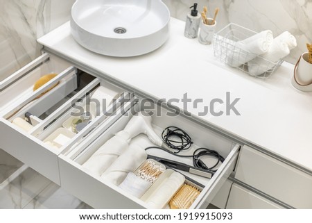 Bathroom under sink organizer drawers with neatly placed bath amenities and toiletries. Table top basin sink with dispenser of liquid soap, toothbrush in cup and group of rolled up white towels.