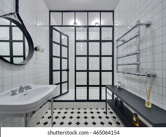 Bathroom With Walls Of White Tiles. There Is White Washbasin, Black Mirror, Shower With Glass Partition With Open Door And Glowing Lamps, Black Rack With Accessories And Chromed Heated Towel Rail. 