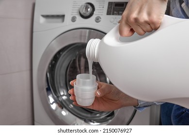 In bathroom where washing machine is located, girl pours the laundry conditioner into lid to give clean laundry pleasant smell and softness. From white plastic bottle, rinse is poured into the cap - Shutterstock ID 2150076927