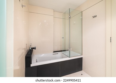 Bathroom with a white bathtub for bathing and relaxation in glossy black marble tiles. Faucet is built into a ledge above the black tile bath. Glass transparent screen protects from splashes. - Powered by Shutterstock