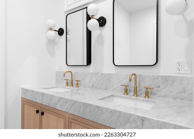 A bathroom with a white oak cabinet, marble countertop, gold faucets, black and white tiled floor, and sconces around black mirrors. - Shutterstock ID 2263304727