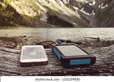 Battery Pack With Solar Battery Charges The Smartphone In The Wild On A Background Of Mountains And Rivers