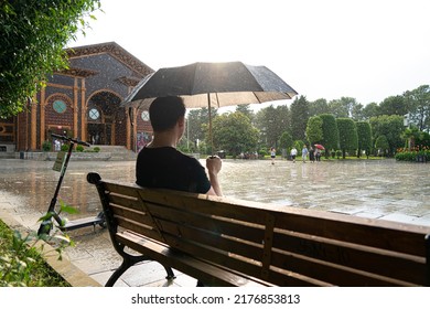 Batumi, Georgia - July 10, 2022: A Man Sits Under An Umbrella On A Bench In The Rain