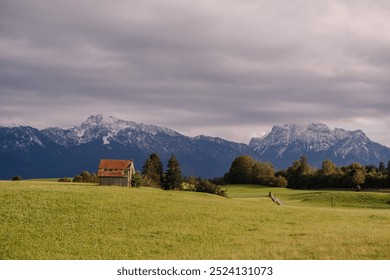 Bavaria, Germany - September 15, 2024 - A small wooden barn stands on a lush green field with snow-capped mountains in the background under a cloudy sky, creating a rural alpine scene. - Powered by Shutterstock