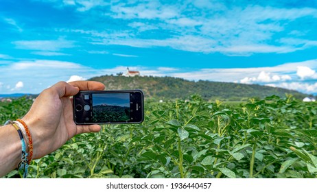 Bavaria, Germany-July 2018: 
Taking A Photo With An IPhone Of A Beautiful Sunny Day Scenery With Beautiful Lush Green Vegetation And A Small Hill With A Catholic Church On Top In The Background. 