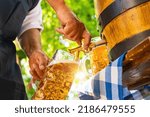 Bavarian man in apron is pouring a large lager beer in tap from wooden beer barrel in the beer garden. Background for Oktoberfest, folk or beer festival (German for: O’zapft is!) 