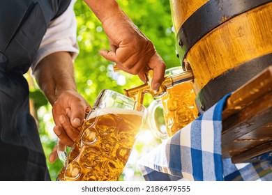 Bavarian man in apron is pouring a large lager beer in tap from wooden beer barrel in the beer garden. Background for Oktoberfest, folk or beer festival (German for: O’zapft is!)  - Shutterstock ID 2186479555
