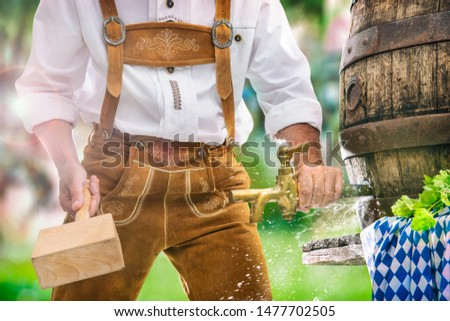 Bavarian man in leather trousers taps a wooden barrel of beer in the garden. Background for Oktoberfest, folk or beer festival 