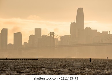 Bay Bridge And Downtown District On Coastal Seafront Of San Francisco. Famous Tourist Attraction Connecting Urban Town. Scenic View Of City Skyline With Sky In Background During Sunset.